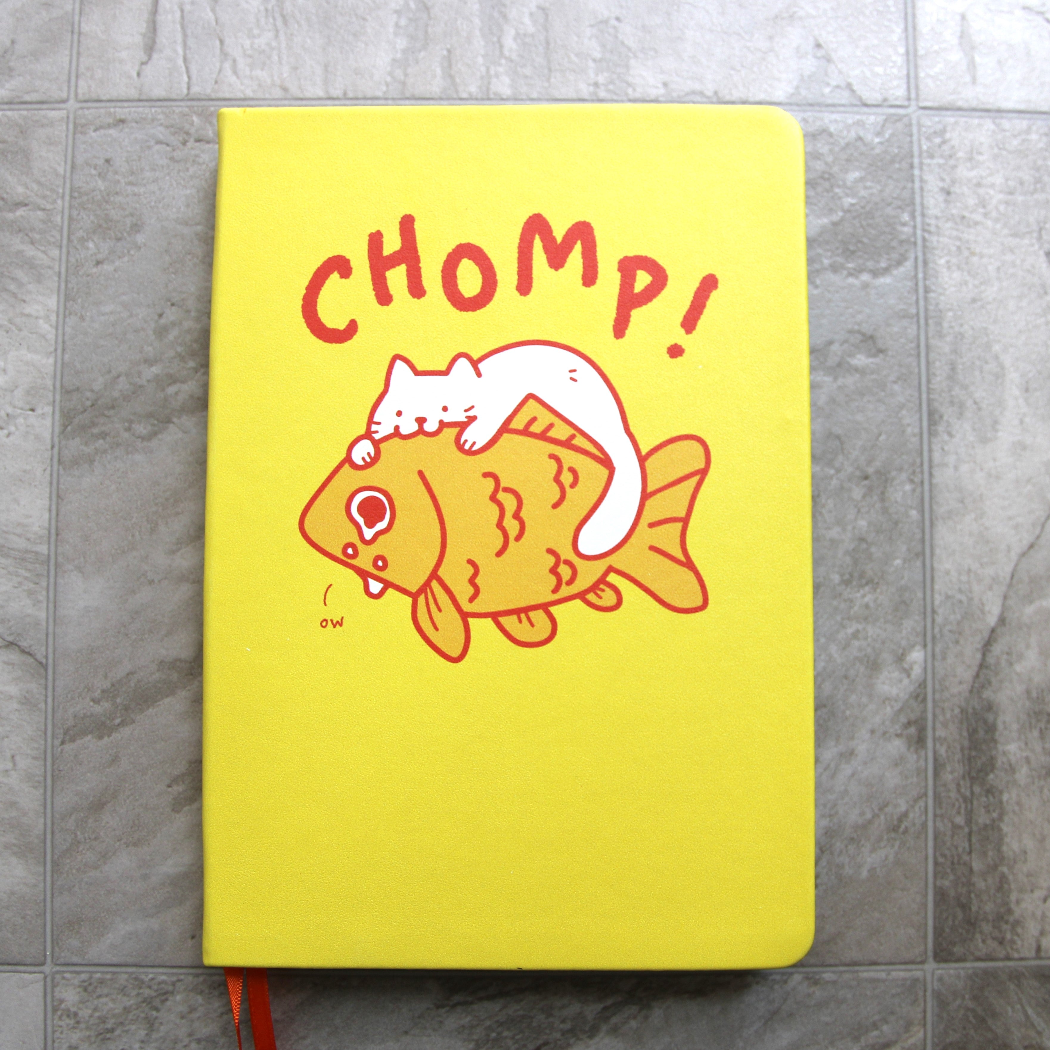 Yellow notebook cover featuring cute artwork of a white cat perched on top of a giant goldfish. The cat is biting the goldfish and the fish is teary-eyed and saying "ow" in small font. Text above art reads "CHOMP!" in large letters