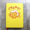 Yellow notebook cover featuring cute artwork of a white cat perched on top of a giant goldfish. The cat is biting the goldfish and the fish is teary-eyed and saying "ow" in small font. Text above art reads "CHOMP!" in large letters