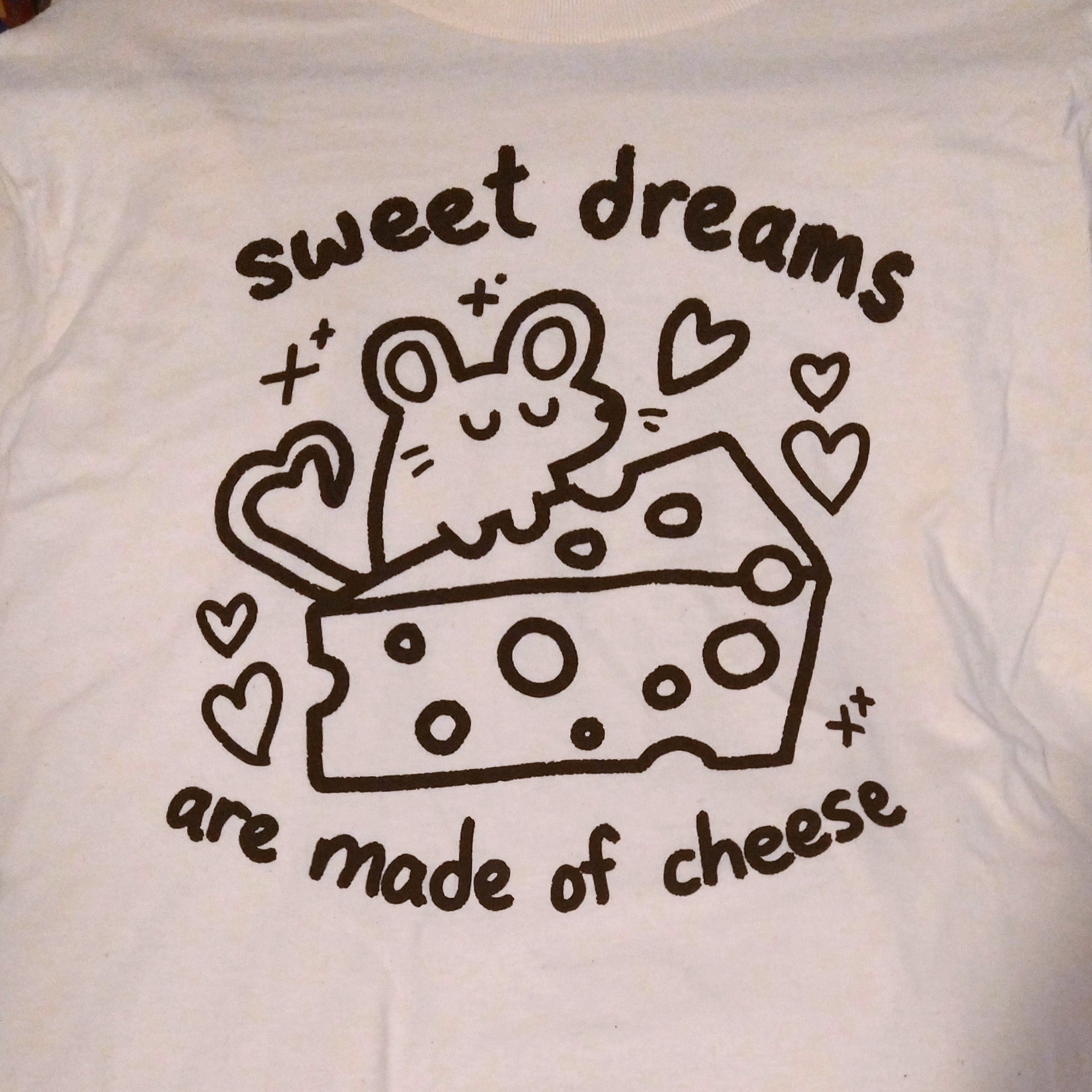 cream colored tee with graphic of a mouse with a block of swiss cheese. the mouse's tail is curled in the shape of a heart. text reads "sweet dreams are made of cheese"
