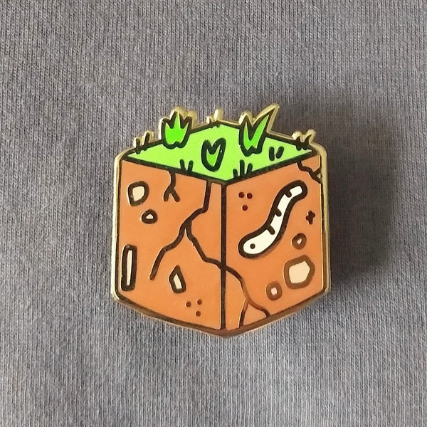 Gold plated hard enamel pin of a cube of dirt with grass on top and a worm