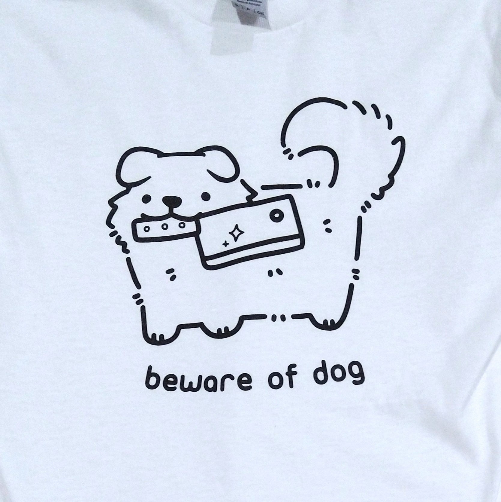 White t shirt printed with black ink graphic of a cute fluffy dog holding a knife (meat cleaver) in its mouth. Text reads "beware of dog"