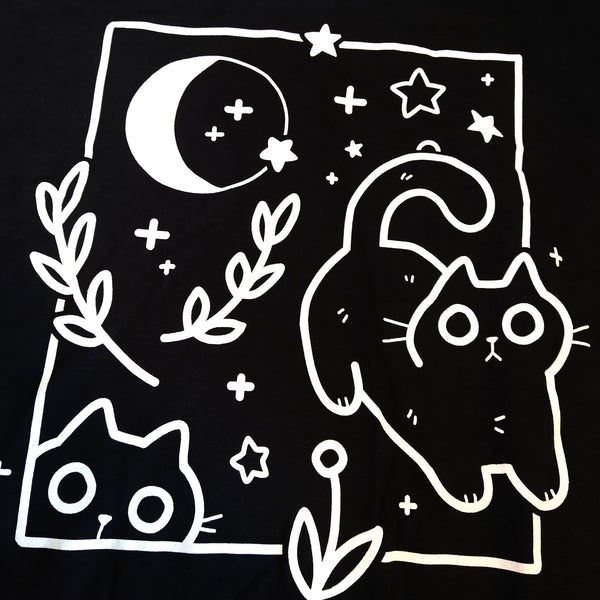 Black tee shirt screen printed with white ink graphic of two black cats surrounded by a rectangular frame, moon, stars, and plants