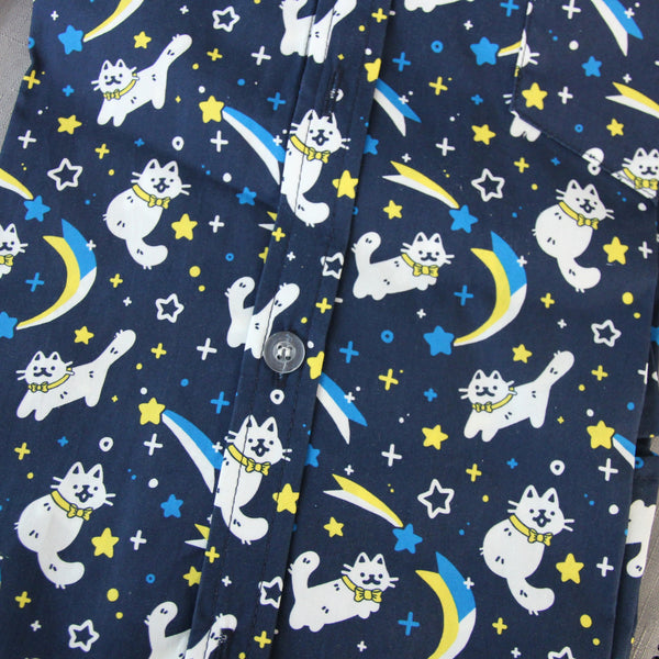 Dark blue button up shirt patterned with white cats wearing yellow bow ties, shooting stars, and sparkles. The cats are drawn in a cute style and the colors of the shirt are white, blue, and yellow.