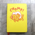 Yellow notebook cover featuring cute artwork of a white cat perched on top of a giant goldfish. The cat is biting the goldfish and the fish is teary-eyed and saying "ow" in small font. Text above art reads "CHOMP!" in large letters