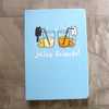 Light blue notebook front cover, featuring artwork of a small black cat drinking from a blue striped straw out of a big glass of lemonade, and a small white cat drinking from a red striped straw out of a glass of orange juice. Each glass has lemon slices and orange slices next to it. Text in white reads "juice friends!"