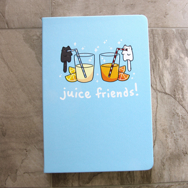 Light blue notebook front cover, featuring artwork of a small black cat drinking from a blue striped straw out of a big glass of lemonade, and a small white cat drinking from a red striped straw out of a glass of orange juice. Each glass has lemon slices and orange slices next to it. Text in white reads "juice friends!"