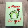 Light green notebook cover featuring a kawaii frog with rosy cheeks holding up a red tulip. Text reads "a flower for you!" in red handwritten lettering
