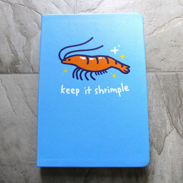 Blue notebook featuring a red shrimp drawing in a cute cartoony style. Text reads "keep it shrimple"