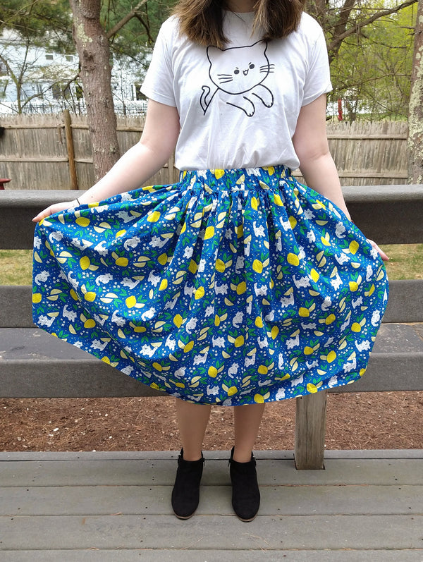 Blue skirt patterned with white cats and lemons. The skirt is midi length and ends below knee.
