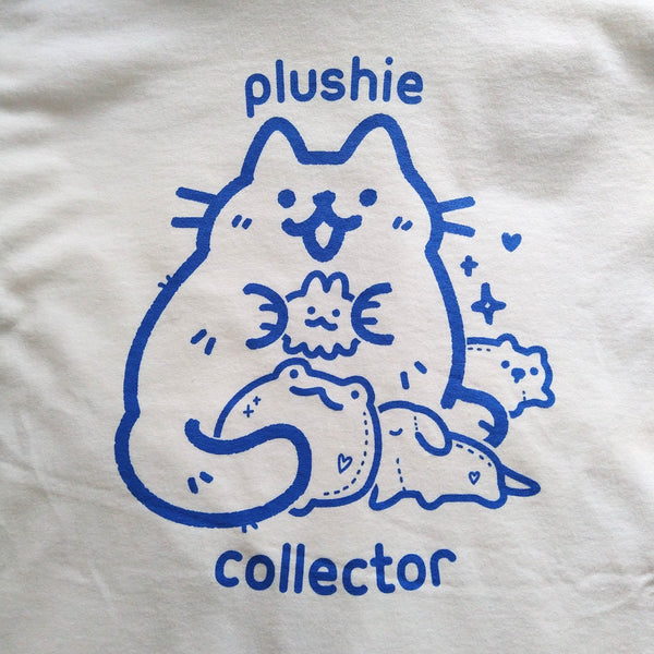 White graphic tee printed with blue ink. Design shows a cute cat holding stuffed animals or plushies. Text "plushie collector"