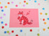 postcard mini print of a red tiger posing cutely and smiling with a pink background