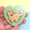 green heart-shaped pinback button with drawing of a cute fluffy yellow dog sticking its tongue out (blep), layered with a holographic heart film