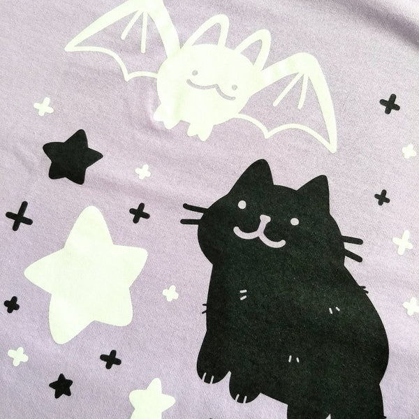 Lavender shirt screen printed with graphic of white bat and black cat drawn in a cute style. They are surrounded by black and white stars and sparkles.