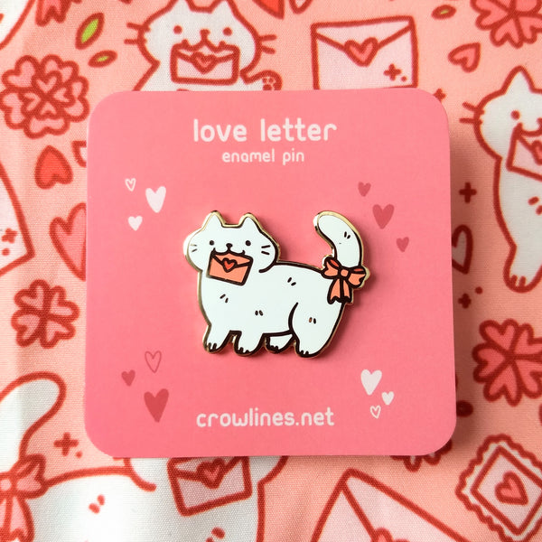 Gold plated hard enamel pin of a white cat holding a pink enveloped sealed with a red heart sticker in its mouth. The cat has a pink ribbon bow on its tail