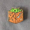 Gold plated hard enamel pin of a cube of dirt with grass on top and a worm