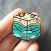 Gold plated hard enamel pin of a cubic beach with waves, a sandy shore, a little crab, starfish, and seashell