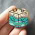 Gold plated hard enamel pin of a cubic beach with waves, a sandy shore, a little crab, starfish, and seashell