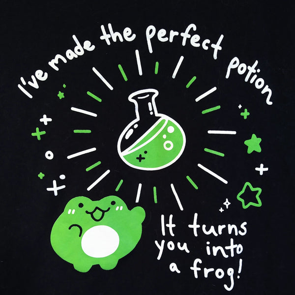 Black tee screen printed with graphic of a frog drawn in a cute style with a round potion flask. Text reads "I've made the perfect potion" "It turns you into a frog!"