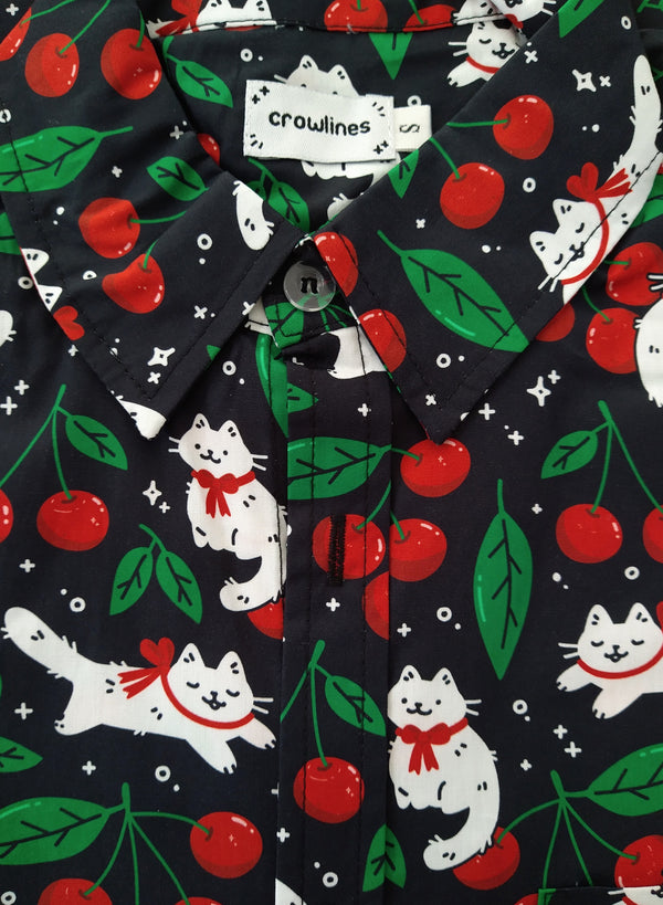 Black button up shirt patterned with cats and cherries. The cats are wearing red ribbons around their necks. The color scheme is black, white, red, and green.