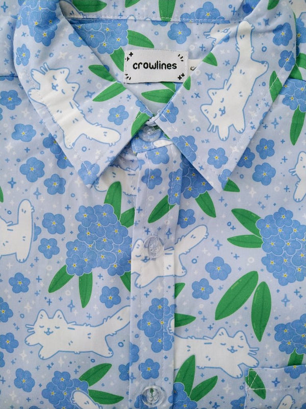 Light blue button up shirt patterned with cats and forget-me-not flowers. The color scheme is blue, white, and green.