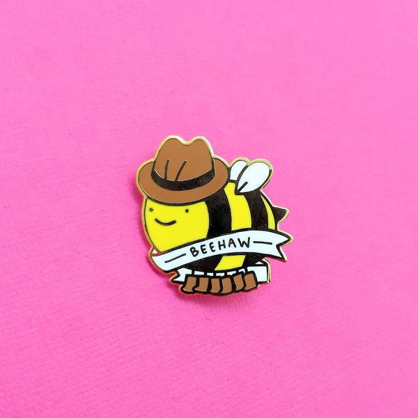 Gold plated hard enamel pin of a bee wearing cowboy hat and cowboy boots with text banner "BEEHAW"